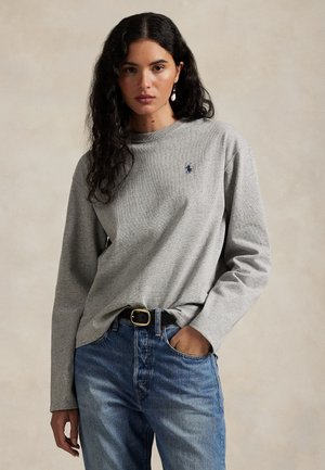 Femme aux longs cheveux foncés portant un t-shirt gris manche longue avec un petit logo et un jean bleu, debout avec une main dans la poche sur un fond uni.