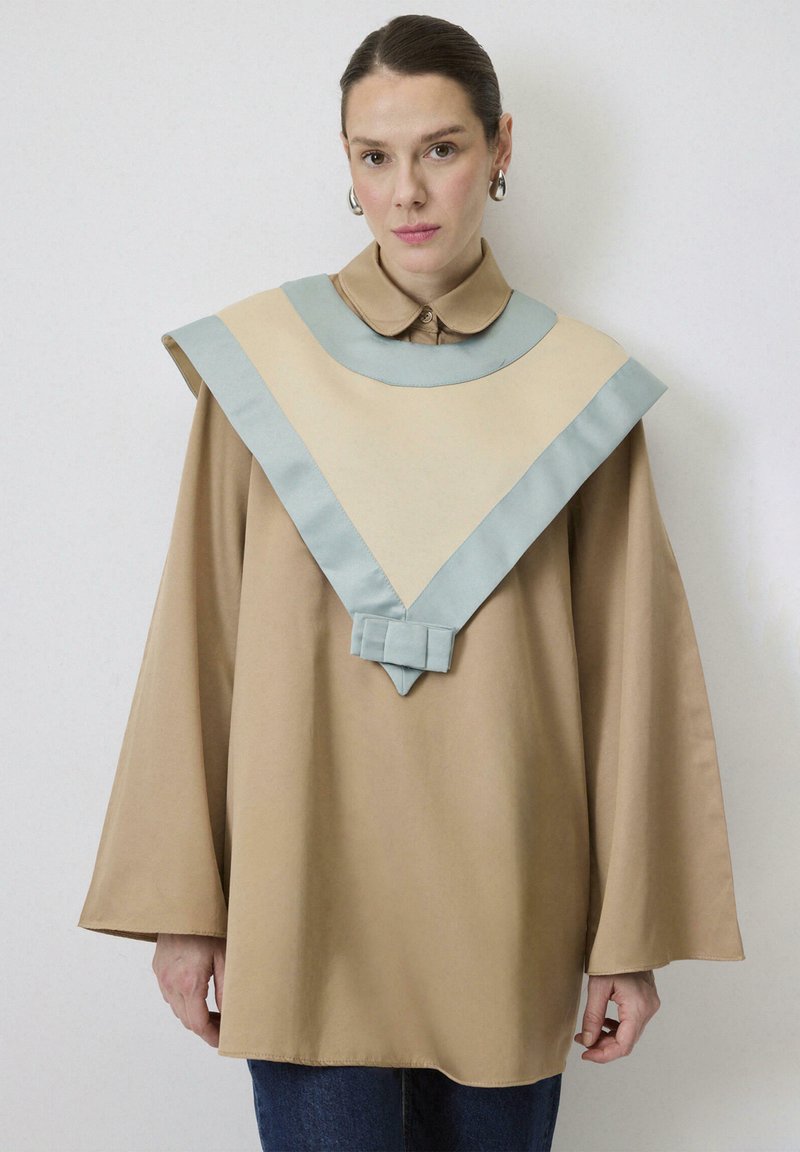 Bežový oversized top s veľkým modrým golierom, ktorý má stredovú mašľu a široké rukávy, vyrobený z hladkej, ľahkej látky.