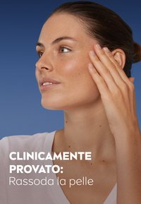 Modella con carnagione media, capelli castano chiaro, che indossa un top bianco a V, che si tocca delicatamente il viso su uno sfondo blu.