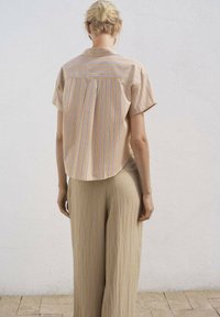 Bluză cu mânecă scurtă, rayată, de culoare piersică, cu dungi albastre și mov, având un tiv curbat și pliu la spate. Asortată cu pantaloni bej deschis, lejeri.