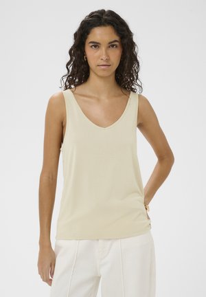 Kobieta o kręconych ciemnych włosach, ubrana w beżowy, bezrękawnik z dekoltem w serek i białe spodnie, stojąca z jedną ręką na biodrze na białym tle.