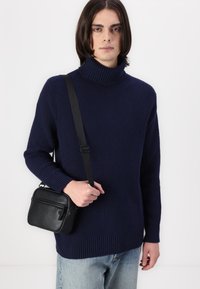Marineblaue gerippte Rollkragenpullover mit legerem Schnitt und langen Ärmeln. In der Hand hält sie eine kleine schwarze Ledertasche mit einem Reißverschlussfach.