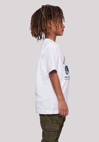 White cotton t-shirt with blue graphic text on the front. The child is wearing olive green cargo pants, showing a relaxed fit and side profile.
