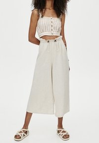 Crème crop top met knopen aan de voorkant en dunne bandjes, gecombineerd met losse, beige wijde broek en beige sandalen met kruislings bandjes.