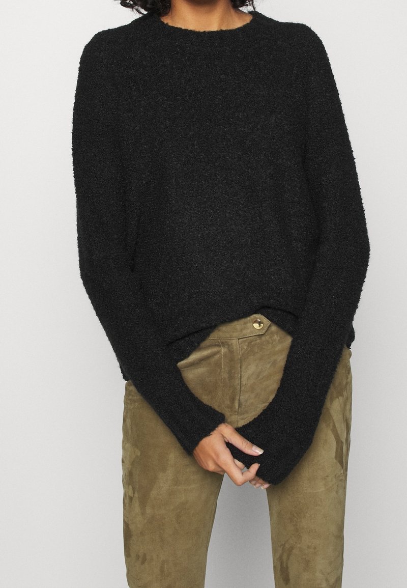 Pull noir et duveteux à manches longues et col rond, associé à un pantalon en daim vert olive avec fermeture à boutons à l'avant.