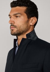 Veste bleu marine avec un col gris texturé, dotée d'une poche poitrine unique et de lignes épurées, associée à une chemise col bleu clair en dessous.