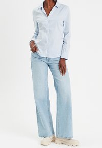 Personne portant une chemise à rayures verticales bleu clair, un jean large délavé, des chaussures beiges épaisses et des bracelets, debout de manière décontractée.