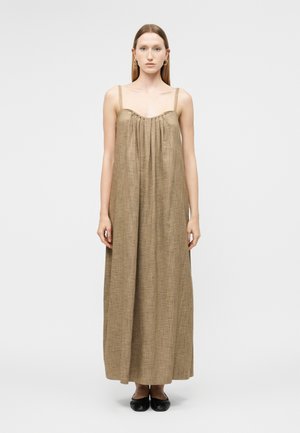 Γυναίκα με ίσια μαλλιά, που φοράει ένα φαρδύ, αμάνικο φόρεμα μήκους μέχρι τους αστραγάλους σε χρώμα ταουπέ και μαύρα μπαλαρίνες, στέκεται μπροστά σε απλό ανοιχτόχρωμο φόντο.