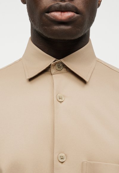 Σκούρου τόνου δέρμα άτομο που φοράει μπεζ πουκάμισο με κουμπιά και μυτερή γιακά, ορατό από το πηγούνι ως το στήθος.