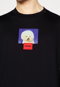 Camiseta negra con un gráfico cuadrado de un perro blanco y esponjoso sobre un fondo azul, con una caja de texto roja etiquetada como "HUGO" debajo.