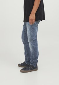 Jeans in denim blu con una vestibilità rilassata, caratterizzati da dettagli strappati, un leggero sbiadimento e accenti cuciti. Abbinati a sneakers scure.