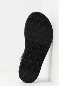 Czarna gumowa podeszwa zewnętrzna z diamentowym wzorem, z wypukłym logo. Cholewka buta w neutralnym kolorze jest częściowo widoczna.