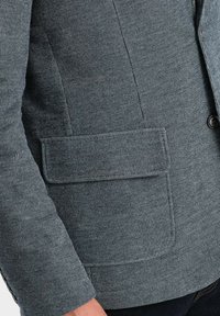 Grå texturerad kavaj med en enkel knappstängning, en ficka med patch och en skräddarsydd passform. Materialet har ett subtilt vävt mönster.