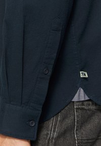 Camicia blu navy con bottoni in tessuto testurizzato, dotata di un orlo arrotondato e una piccola etichetta vicino alla cucitura laterale, con un dettaglio delle maniche con bottoni.