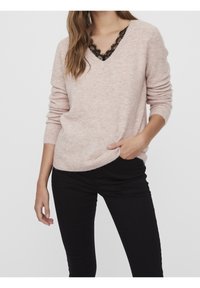 Pull en maille rose clair avec encolure en V et accents en dentelle noire. Manches longues et coupe décontractée, porté avec un pantalon noir. Texture douce.