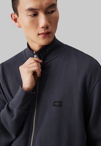 Dunkelgraue Zip-Jacke mit hohem Kragen, strukturiertem Stoff und einem kleinen Logo-Patch. Mit Reißverschluss und gerippten Bündchen.