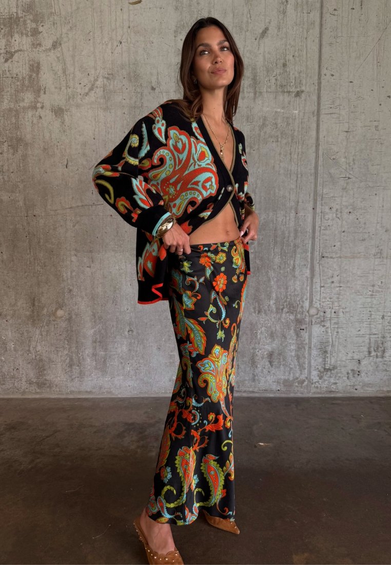 Γυναίκα που φοράει μαύρο ζακέτα και μακριά φούστα με πολύχρωμα σχέδια πεϊσλεϊ, στέκεται μπροστά σε γκρι τσιμεντένιο τοίχο, διορθώνοντας τη μέση της φούστας της.