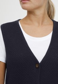 Dunkelblaues Strickwesten mit strukturiertem Muster, V-Ausschnitt und einem einzelnen braunen Knopf. Über einem weißen T-Shirt getragen. Glatte Textur.