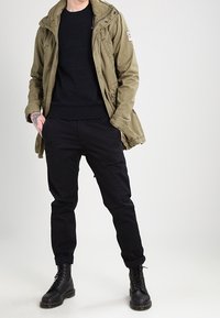 Parka verde oliva con cerniera e bottoni, maglione nero, pantaloni neri e stivali neri stringati con superficie strutturata.