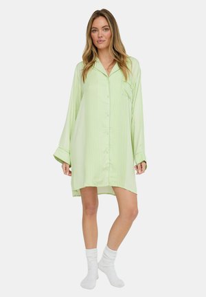 Frau trägt ein hellgrün gestreiftes, übergroßes Hemdblusenkleid und weiße Socken, steht vor einem schlichten hellen Hintergrund.