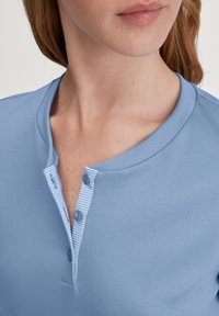 Hellblaues Henley-Shirt mit geripptem Kragen, gestreiftem Knopfverschluss und zwei Knöpfen. Der Stoff wirkt weich mit einer glatten Textur.