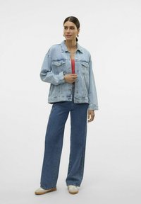 Zbledečena modra denim jakna z velikimi sprednjimi žepi in gumbom za zapiranje, v kombinaciji z širokimi modrimi kavbojkami in rdečo majico, prikazana na modelu.