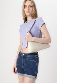 Svetlofialové skrátené tričko s ryhovanou textúrou, tmavomodré džínsy s ohrnutými okrajmi a béžová kabelka Calvin Klein s hladkým povrchom.