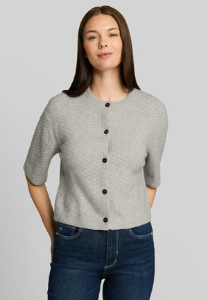 Femme aux cheveux bruns et lisses portant un pull gris clair à manches courtes boutonné et un jean bleu foncé, debout devant un fond gris uni.