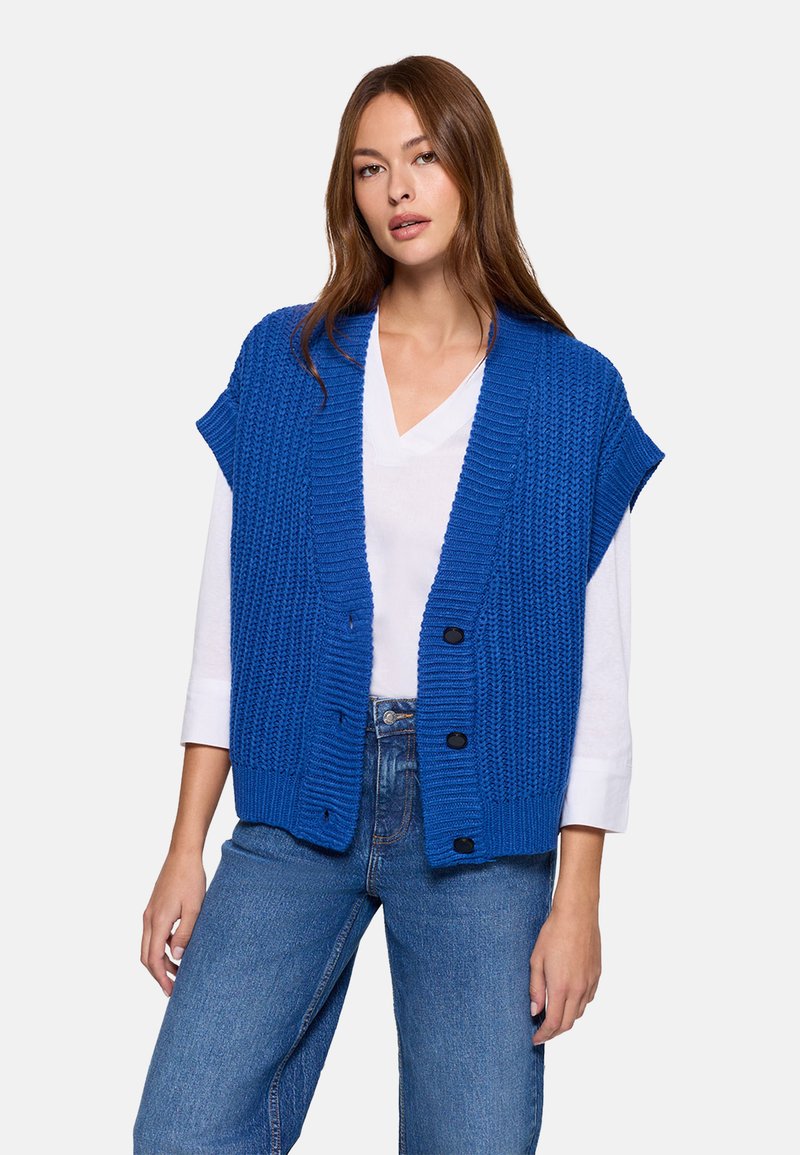 Chaleco de punto azul con textura acanalada, abertura frontal y mangas cortas, con botones negros y llevado sobre una camisa blanca.