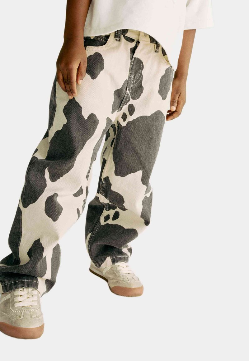 Pantaloni in denim con stampa zebrata bianca e nera, dal taglio morbido, abbinati a sneaker grigie chiare e una camicia bianca. Focus su uno stile casual e un motivo unico.