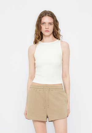 Jeune femme aux cheveux ondulés portant un crop top blanc sans manches et un short beige à cordon, debout devant un fond clair uni.