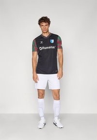 Homme portant un maillot de football noir avec des manches à motifs rouges et verts, un short blanc, de longues chaussettes blanches et des chaussures à crampons blanches, debout devant un fond uni.