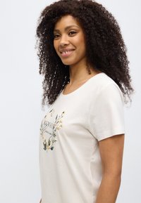 Μπεζ μπλουζάκι με κοντά μανίκια, με floral σχέδιο και το κείμενο «Always Flourishing» σε διακοσμητική γραμματοσειρά στο μπροστινό μέρος.