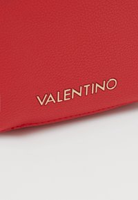 Röd läderväska med texturerad yta, präglad "Valentino"-logotyp i guld och precist sytt längs kanterna.