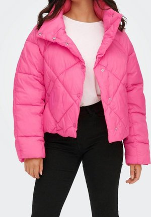 Vrouw draagt een felroze gewatteerd donsjack over een wit shirt en zwarte broek, staand tegen een effen witte achtergrond.