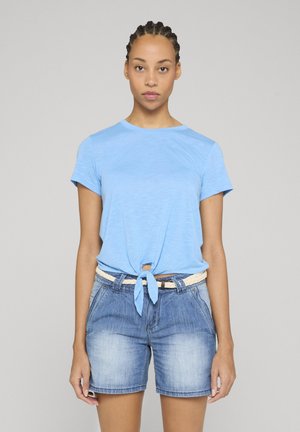 Giovane donna con capelli intrecciati che indossa una camicia azzurra a maniche corte annodata e pantaloncini di jeans con una cintura bianca intrecciata, in piedi immobile.