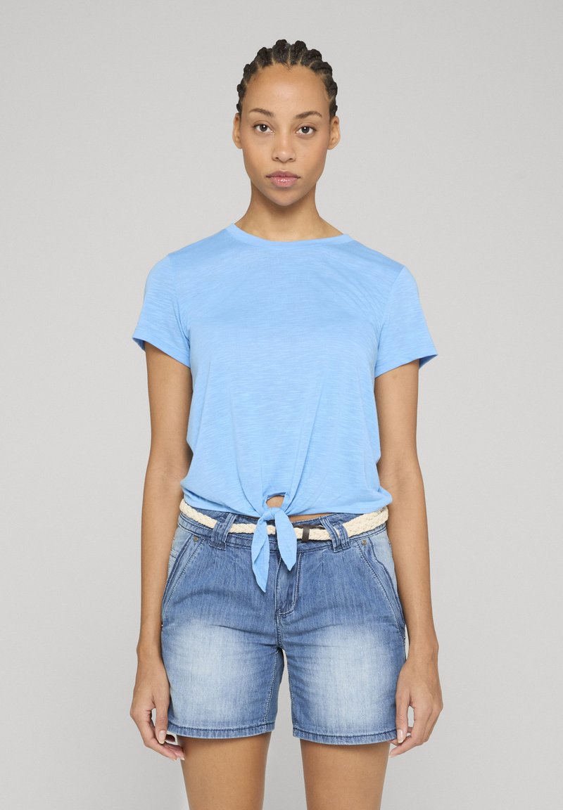 Jeune femme aux cheveux tressés portant une chemise nouée à manches courtes bleu clair et un short en jean avec une ceinture blanche tressée, debout immobile.