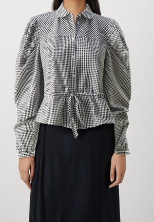 Žena v černobílé kostkované blůze s nabíranými dlouhými rukávy a zavázaným pasem, v kombinaci s černou sukní.