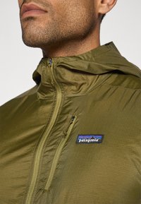 Patagonia lightweight jacket in olive green, featuring a zip closure, hood, visible texture, and logo patch on the chest.