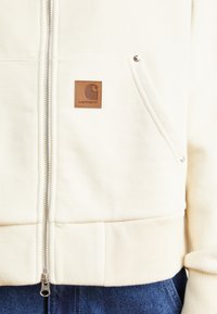 Light cream zip-up sweatshirt with ribbed cuffs, side pocket, and a tan leather logo patch on the chest. Smooth fabric texture.
