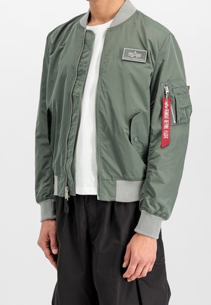 Mand iført grøn Alpha Industries bomberjakke med ribbede manchetter og krave, lynlås foran og rød "Remove Before Flight"-mærkat på ærmelommen.