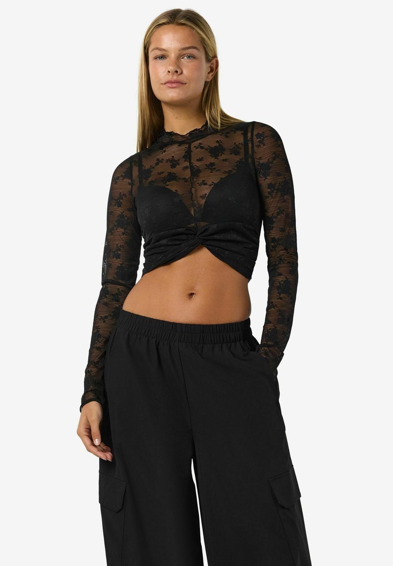 Fekete áttetsző, hosszú ujjú felső virágos csipke mintázattal, csavart elülső résszel. Lazán illeszkedő fekete cargo nadrággal párosítva, oldalsó zsebekkel.