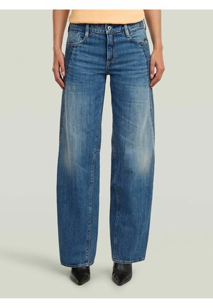 Person, die locker sitzende blaue Jeans und schwarze spitze Schuhe trägt, steht vor einem einfarbigen hellen Hintergrund.