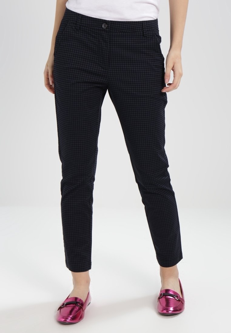 Femme portant un pantalon slim noir à carreaux associé à des mocassins métalliques roses et un haut blanc, debout devant un fond uni.