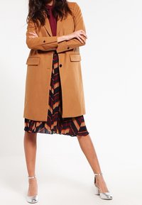 Manteau couleur chamel avec des épaules structurées et deux poches avant, superposé à une robe plissée à rayures multicolores et des talons argentés.