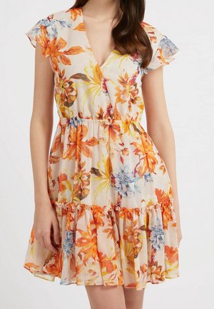 Vrouw draagt een korte mouwloze bloemenjurk met oranje, gele en blauwe bloemen en een getailleerde taille met een ruches zoom.