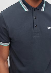 Mörkblå polo skjorta med tre svarta knappar och randig krage och ärmar, bärs av en person med medelbrun hudton.