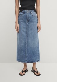 Kvinna iklädd en mid-blå denim midi kjol, svart ärmlös topp och svarta sandaler med öppen tå som står mot en enkel bakgrund.