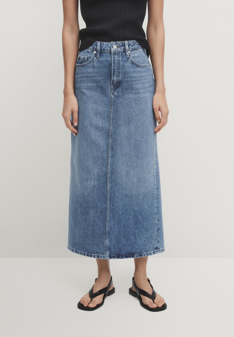 Kvinna iklädd en mid-blå denim midi kjol, svart ärmlös topp och svarta sandaler med öppen tå som står mot en enkel bakgrund.