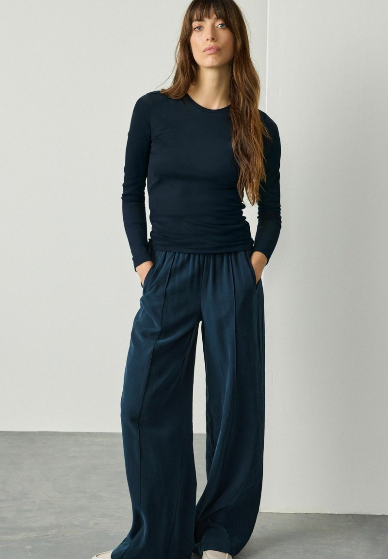 Vrouw met lang bruin haar, gekleed in een nauwsluitende zwarte top met lange mouwen en een brede broek in marineblauw, staande met haar handen in de zakken tegen een effen muur.